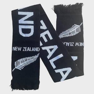 May include: A black knit scarf featuring white text that reads NEW ZEALAND and a white fern leaf graphic. The scarf has black fringe at both ends and displays the word ZEALAND in large white letters running diagonally across the fabric.