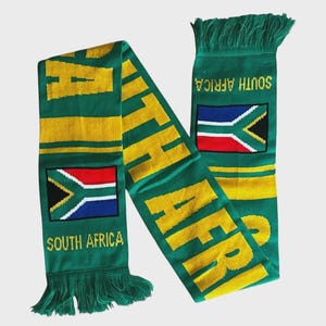 May include: A green knit scarf featuring the text SOUTH AFRICA in yellow letters, along with the South African national flag. The scarf includes yellow horizontal stripes and green fringe at both ends, displaying a bold, colourful design.