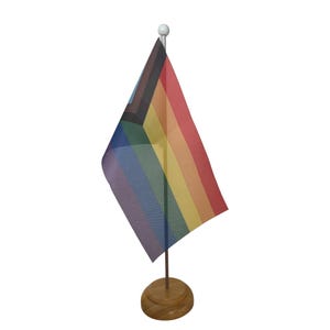 May include: A rainbow pride flag on a wooden desk stand. The flag has six stripes: black, brown, light blue, dark blue, light pink, and yellow.