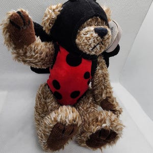 May include: A plush teddy bear dressed as a ladybug. The bear has brown fur, a black hood, and a red vest with black spots. It has small black wings and is posed on a white surface.