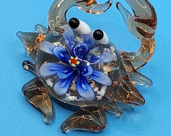 May include: A glass crab figurine with a blue flower design. The crab has clear and brown accents, with black eyes and a blue flower with a yellow center. The figurine is set against a blue background.