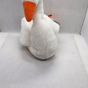 Può includere: Un giocattolo di peluche bianco a forma di oca con becco arancione e occhi neri. L'animale di peluche è fatto di materiale morbido e soffice ed è seduto. Il giocattolo ha un'etichetta.