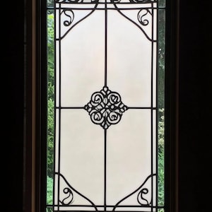 May include: A window with a stained glass design featuring a black floral pattern on a white background. The window is framed in dark wood and has a view of green foliage outside.