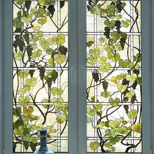 Puede incluir: Una ventana de doble panel con vidrieras de motivo de vid, que muestra hojas verdes y racimos de uvas de color púrpura oscuro sobre un fondo claro. Un jarrón de cerámica azul y blanco se asienta en el alféizar frente a la ventana.