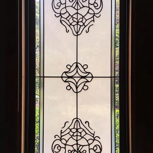 May include: A window with a frosted glass pane featuring a decorative black iron design. The design is made up of three repeating patterns of swirls and curves.