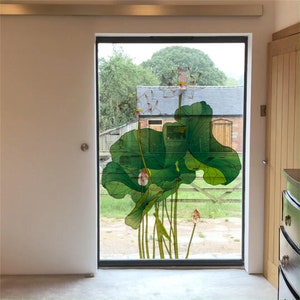 Könnte beinhalten: Ein Fenster mit einer dekorativen Folie, die ein großes grünes Lotusblüten-Design zeigt. Die Blume blüht mit rosa und weißen Blütenblättern. Das Fenster ist schwarz gerahmt und in eine weiße Wand eingefasst.