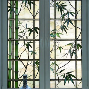 Puede incluir: Una ventana con marco blanco con un diseño de vidriera que presenta tallos y hojas de bambú verdes. La ventana está dividida en secciones rectangulares. Un jarrón con estampado azul y blanco se encuentra en el alféizar.