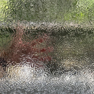 Pode incluir: Uma imagem em close-up de uma superfície de vidro texturizada com um padrão repetitivo. O vidro transparente permite vislumbrar cores verdes e vermelhas desfocadas atrás. O padrão cria um efeito distorcido semelhante a um mosaico.