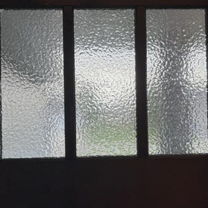 Puede incluir: Una ventana de tres paneles con vidrio texturizado. El vidrio tiene una superficie ondulada e irregular, creando una vista distorsionada. El marco de la ventana es de color marrón oscuro y el fondo es ligeramente visible a través del vidrio.