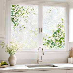 Puede incluir: Una película para ventanas con un patrón de flores blancas y hojas verdes aplicada en una ventana de dos paneles. El diseño incluye racimos de pequeñas flores blancas y follaje verde que se extienden desde las esquinas de los cristales.