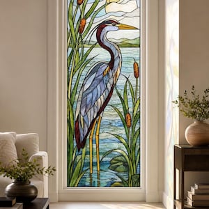 May include: A stained glass window panel featuring a tall heron standing in water among green cattails and lily pads. The background depicts a lake, distant hills, and a pale sun in the sky. The glass uses shades of blue, green, brown, and yellow.