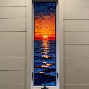 May include: Stained glass window depicting a sunset over the ocean. The sky transitions from blue to orange, with a large sun setting over the water. The ocean is a deep blue with shimmering reflections. The window is framed in white.