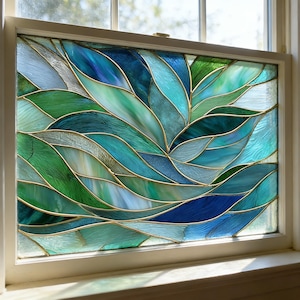 May include: Stained glass window panel with a leaf-like design. The panel features shades of blue and green, with gold-coloured metal lines separating the glass pieces. The window is set in a white frame, creating a decorative element.
