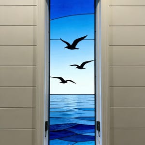 May include: A stained glass window depicting a seascape with three black seagulls in flight against a blue sky. The lower portion shows the ocean in shades of blue. The window is framed in white and set in a white paneled wall.