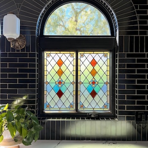 Op de afbeelding: Een gebrandschilderd raam met een zwart frame, met een ruitpatroon in tinten rood, oranje, blauw en groen. Het raam is geplaatst in een zwarte, gewelfde bakstenen muur. Een witte lamp en een potplant zijn ook zichtbaar.