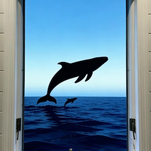 Könnte beinhalten: Ein Fenster mit Blick auf das Meer und zwei schwarzen Silhouetten von Delfinen, die aus dem Wasser springen. Der Himmel ist ein Blauton, und der Fensterrahmen ist weiß.