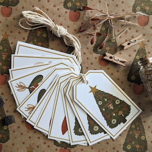 May include: A set of ten white gift tags with gold trim, featuring a hand-drawn illustration of a Christmas tree decorated with ornaments, mushrooms, and a star. The tags are tied together with twine.