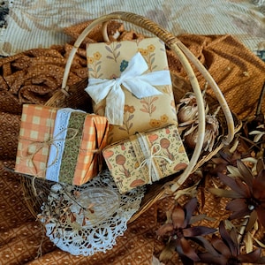 May include: Three rectangular gift boxes wrapped in patterned paper inside a woven wicker basket. One box features a white ribbon, another has orange gingham and green floral fabric with lace, and the third displays a mushroom print with twine.