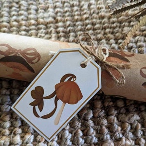May include: A white gift tag with a brown mushroom illustration and a brown bow. The tag is tied with twine and is on a brown and beige woven rug.