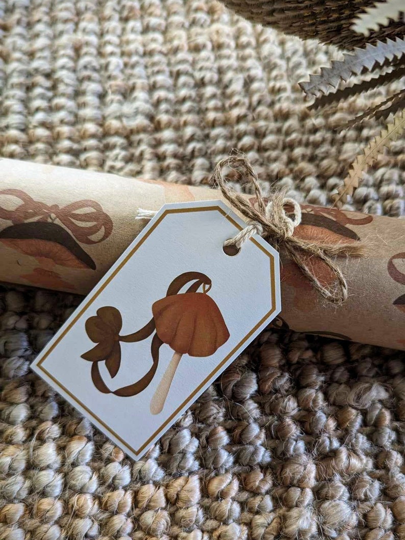 May include: A white gift tag with a brown mushroom illustration and a brown bow. The tag is tied with twine and is on a brown and beige woven rug.