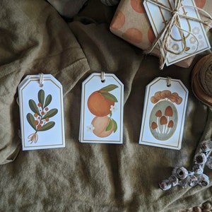 May include: Three white gift tags with different illustrations. The first tag features a green plant with red berries. The second tag has a drawing of two orange fruits with white flowers. The third tag shows a drawing of mushrooms in a frame.