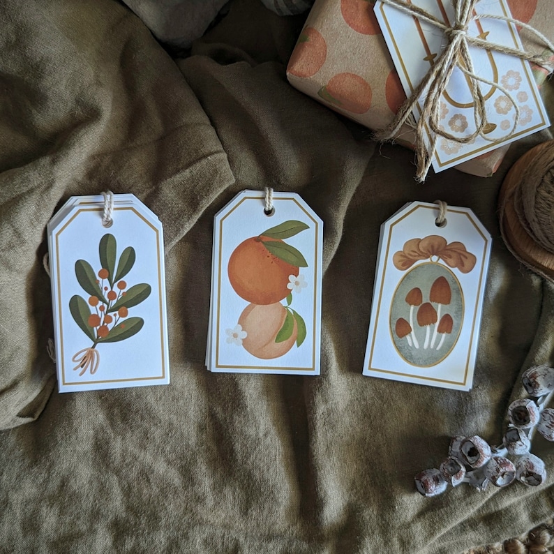 May include: Three white gift tags with different illustrations. The first tag features a green plant with red berries. The second tag has a drawing of two orange fruits with white flowers. The third tag shows a drawing of mushrooms in a frame.
