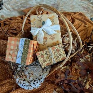 Puede incluir: Tres cajas de regalo rectangulares envueltas en papel estampado y colocadas dentro de una cesta de mimbre tejida. Una caja presenta un estampado floral amarillo con una cinta blanca, otra un patr&oacute;n de cuadros naranja con encaje y la tercera un estampado de setas.