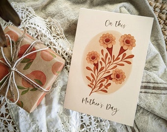 May include: A card with a floral illustration of orange flowers and leaves on a peach background. The text "On this Mother's Day" is printed on the card.