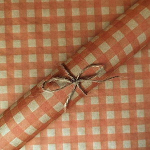 Puede incluir: Un rollo de papel de regalo con un patr&oacute;n de cuadros vichy en naranja y beige. El papel est&aacute; sujeto con un cordel de yute marr&oacute;n natural fino atado en un lazo sencillo alrededor del centro del rollo, sobre una hoja de papel a juego.