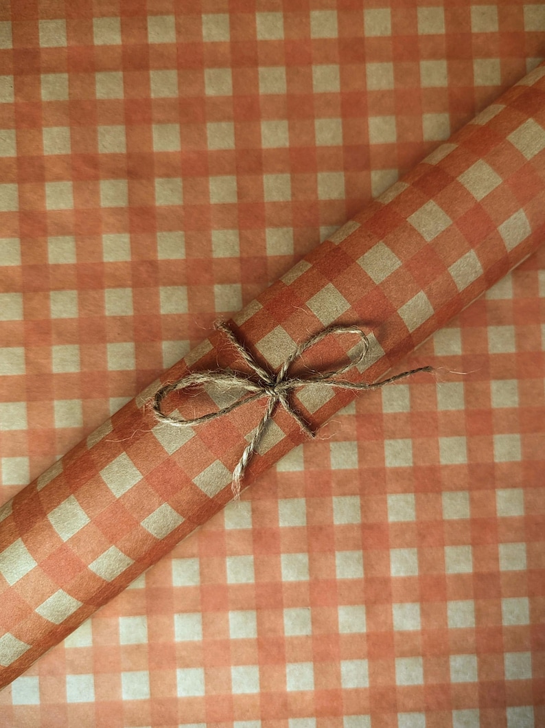 Puede incluir: Un rollo de papel de regalo con un patr&oacute;n de cuadros vichy en naranja y beige. El papel est&aacute; sujeto con un cordel de yute marr&oacute;n natural fino atado en un lazo sencillo alrededor del centro del rollo, sobre una hoja de papel a juego.