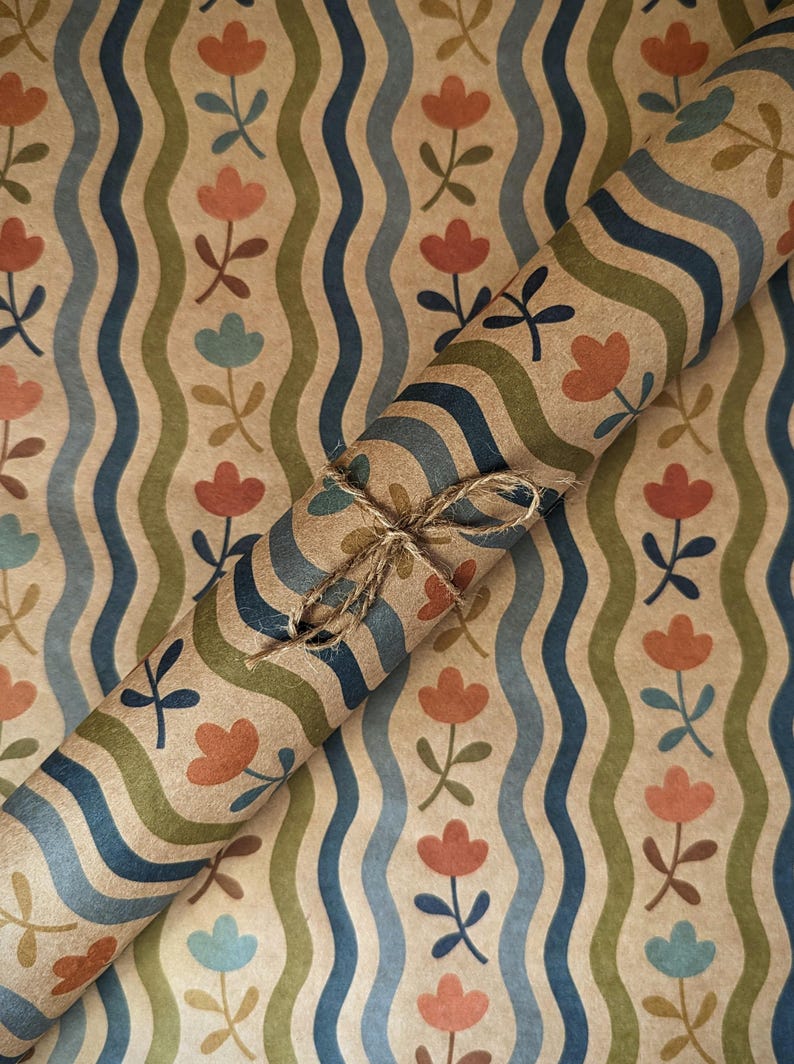 Puede incluir: Un rollo de papel de regalo kraft marr&oacute;n con un patr&oacute;n repetitivo de l&iacute;neas onduladas verticales en azul oscuro y verde oliva, intercaladas con peque&ntilde;as flores naranjas y azul claro, atado con un fino cordel de yute natural.