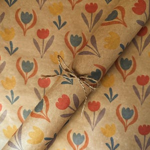 Puede incluir: Un rollo de papel de regalo kraft marr&oacute;n con un patr&oacute;n repetitivo de tulipanes estilizados en tonos rojo, amarillo y azul apagado. El papel est&aacute; atado con un trozo fino de cuerda de yute natural envuelto alrededor del centro del rollo.