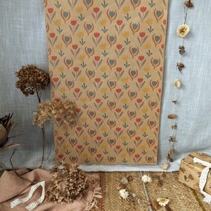 Puede incluir: Una hoja rectangular de papel kraft marr&oacute;n con un patr&oacute;n repetitivo de tulipanes estilizados en tonos rojo, amarillo y azul gris&aacute;ceo apagado. El papel se muestra verticalmente, exhibiendo un estampado floral consistente en toda su superficie.