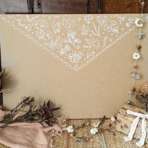 Puede incluir: Un sobre grande de cart&oacute;n color canela con un patr&oacute;n floral blanco dibujado a mano en el borde de la solapa superior. El sobre est&aacute; decorado con flores secas, una peque&ntilde;a caja de regalo con cinta de encaje blanca y una tela rosa claro.
