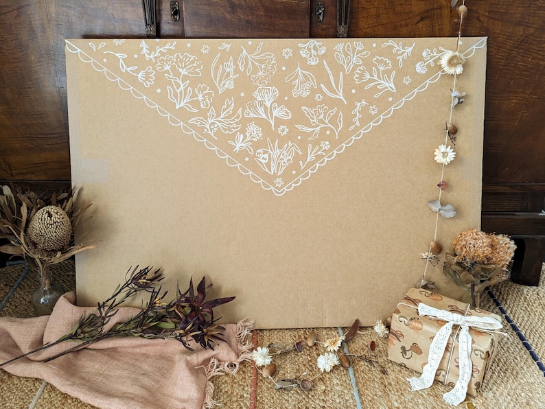 Puede incluir: Un sobre grande de cart&oacute;n color canela con un patr&oacute;n floral blanco dibujado a mano en el borde de la solapa superior. El sobre est&aacute; decorado con flores secas, una peque&ntilde;a caja de regalo con cinta de encaje blanca y una tela rosa claro.