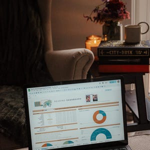 May include: A laptop displaying a "Reading Dashboard" spreadsheet with charts and data. The laptop is open on a couch, near a chair, books, a candle, and a mug. The screen shows data visualization.