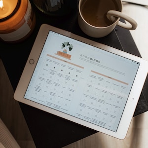 May include: A digital tablet displaying a "Book Bingo" game, with a cup of coffee and a lit candle in the background. The bingo card features various reading challenges. The tablet is on a dark surface, with a beige mug and a candle in a glass jar.