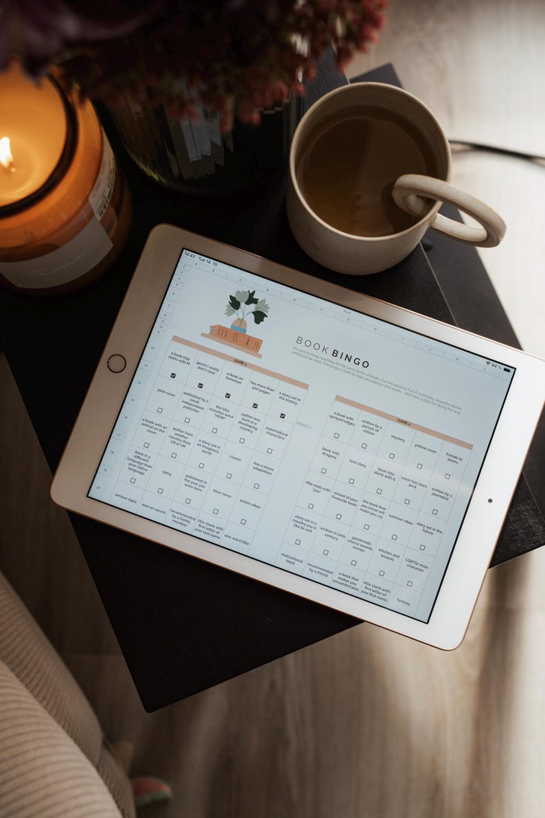 May include: A digital tablet displaying a "Book Bingo" game, with a cup of coffee and a lit candle in the background. The bingo card features various reading challenges. The tablet is on a dark surface, with a beige mug and a candle in a glass jar.