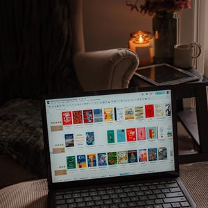 May include: A laptop displaying a digital reading tracker with book covers. The laptop sits on a couch next to a side table with a candle, flowers, and a mug. The scene is dimly lit, creating a cozy atmosphere.