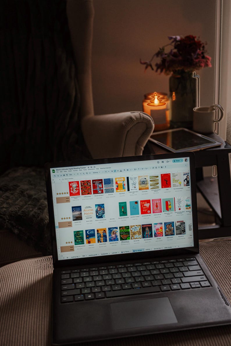 May include: A laptop displaying a digital reading tracker with book covers. The laptop sits on a couch next to a side table with a candle, flowers, and a mug. The scene is dimly lit, creating a cozy atmosphere.