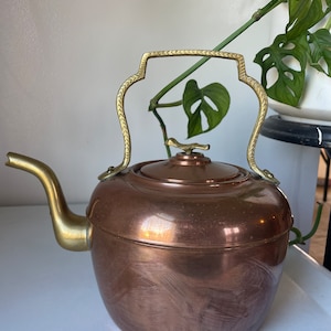 Könnte beinhalten: Ein Kupfer-Teekessel mit einem Messinggriff und Ausguss. Der Kessel hat eine glänzende, polierte Oberfläche und ist in gutem Zustand.