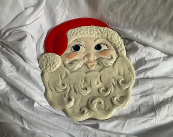 May include: A ceramic Santa Claus face with a red hat, white beard, and rosy cheeks. The Santa face is a decorative item, likely for Christmas. The eyes are blue, and the beard has a textured, curly design.