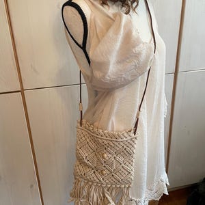 May include: A beige crocheted crossbody bag with fringe detailing and wooden beads. The bag is worn on a mannequin wearing a white dress.