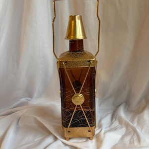 May include: A vintage amber glass bottle with a gold-colored metal frame and handle. The bottle has a decorative design and a gold-colored cap. The metal frame has a geometric pattern. The bottle is rectangular and has a unique, antique appearance.