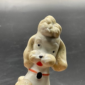 May include: A white porcelain figurine of a poodle with a red and black collar. The poodle has a fluffy white coat and black eyes.