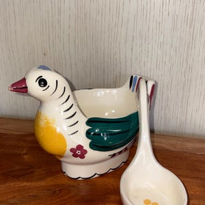 May include: A cream-coloured ceramic bird-shaped bowl featuring a yellow chest, green wings, and a small red flower detail. The bird has a dark red beak and black eye markings. A matching cream-coloured ladle with a small yellow flower design is included.