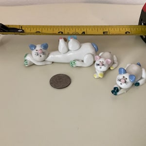 May include: Three white ceramic cat figurines with blue and green accents. The cats are in various poses, including lying down and sitting up. The figurines are about 2 inches tall.
