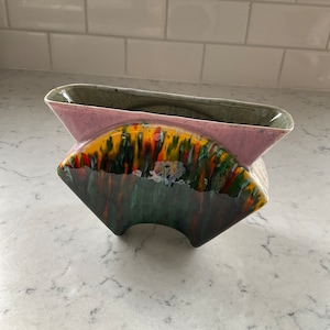 May include: A ceramic planter with a unique abstract design. The planter is a light pink color with green, yellow, orange, and black accents. The planter has a wide, open top and a rounded base.