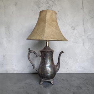 Może przedstawiać: Lampa w stylu vintage z podstawą w kształcie srebrnego czajnika i beżowym abażurem. Czajnik ma zakrzywiony uchwyt i dzióbek i stoi na trzech małych nóżkach. Abażur jest w jasnobrązowym kolorze i jest lekko zwężony.