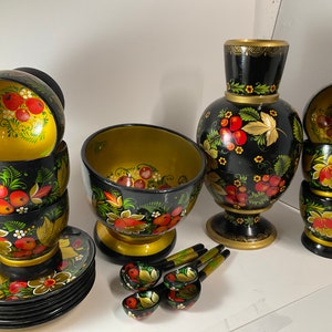 May include: A set of hand-painted wooden bowls, cups, and a pitcher with a floral design. The set features black, gold, red, and green colors. The bowls and cups are stacked, and the pitcher is on a stand. There are also four wooden spoons with floral designs.